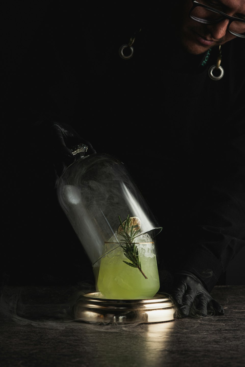 Bartender lifting a glass cloche from a garnished green cocktail.