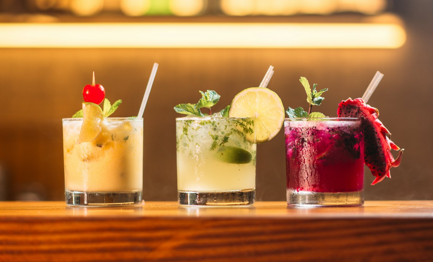 Three colorful cocktails garnished with fruit on a wooden bar.