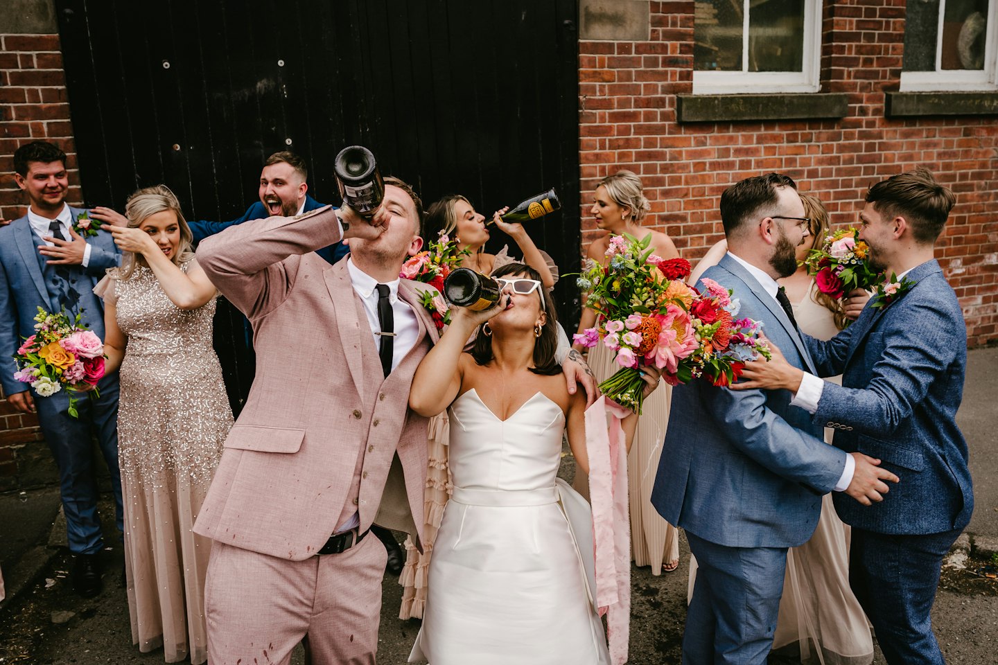 Wedding guests celebrate while drinking champagne outside a brick building.
